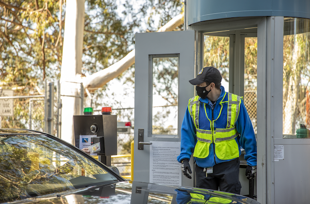 A security guard in a blue shirt and neon safety vest leans out of a guard booth to speak to the driver of a car entering through the Lab's secured gate