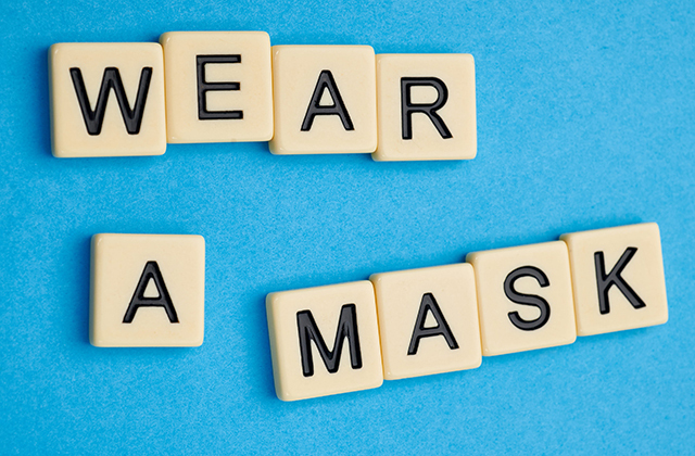 the words "wear a mask" are spelled out on tan Scrabble tiles (with black letters) against a blue background