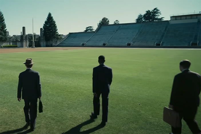 3 men in suits and carry briefcases walking across a football field