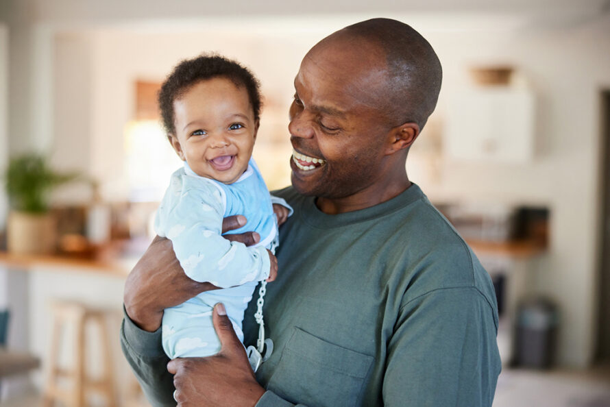 while standing with his laughing little baby baby in his arms at home in their living room