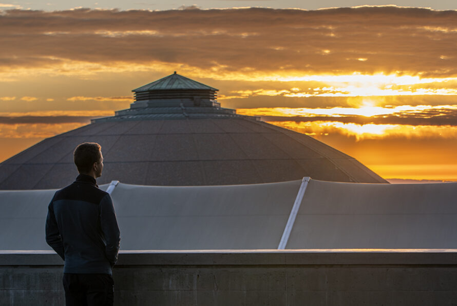 Person looking toward horizon at sunset with large domed building in background.