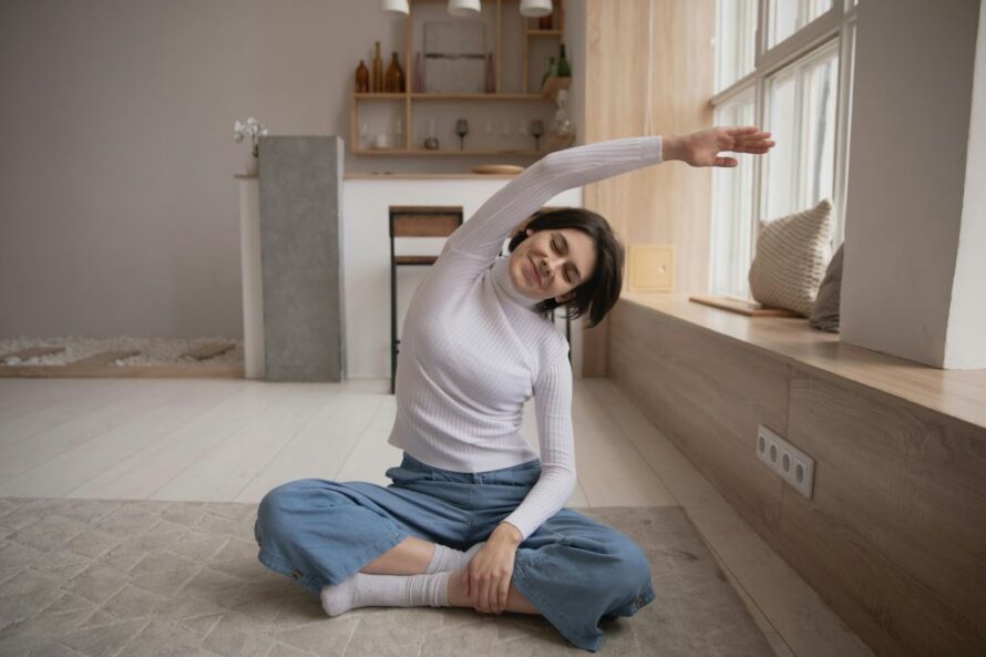 A person sits on the floor in a comfortable, cross-legged position, performing a gentle side stretch with one arm overhead. The scene takes place in a cozy, minimalistic room with large windows, soft natural light, and warm-toned decor.