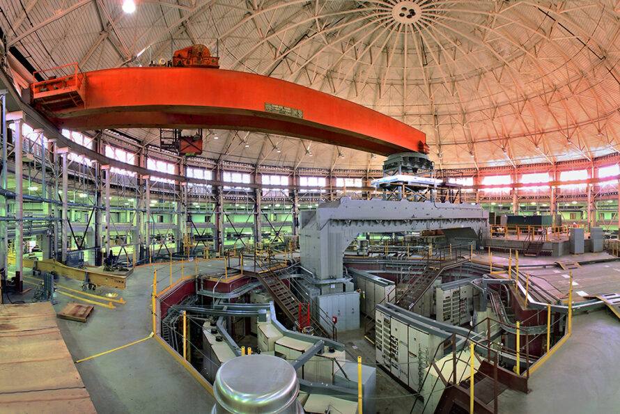 Interior of a large industrial facility with a domed ceiling, featuring a prominent orange overhead crane, structural beams, and various machinery and equipment used for scientific research or engineering purposes.