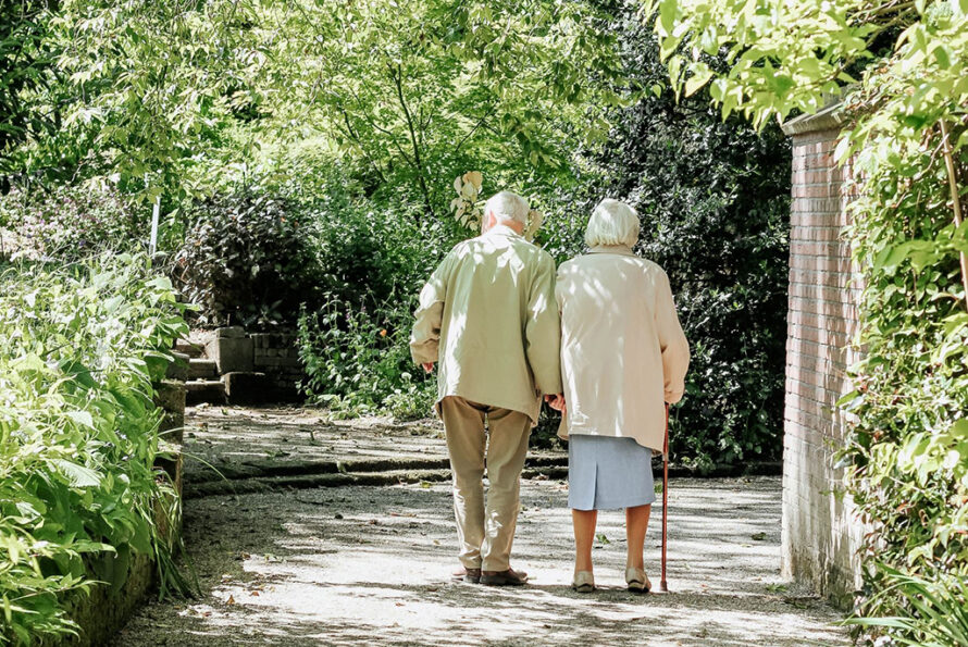 Two elderly people walking hand in hand down a shaded garden path, one using a cane, symbolizing care and companionship in old age.