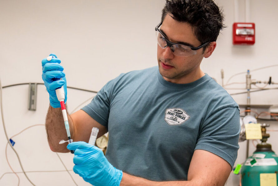 Researcher wearing safety goggles and gloves uses a pipette in a laboratory setting, focusing on a sample in a test tube