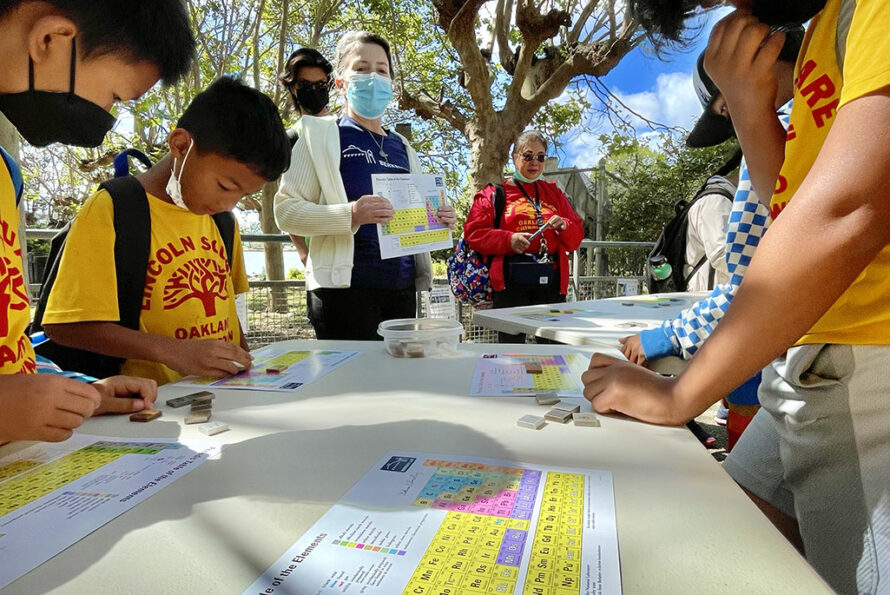 Students participating in hands-on activities at a science fair, with volunteers assisting them in exploring the periodic table and educational experiments