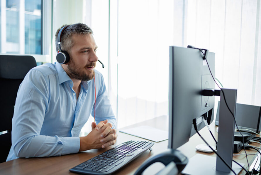 Man using electronic technology at work, attending video conference with headphones on, looking at computer monitor