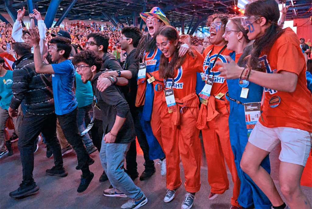 A group of excited participants in colorful uniforms cheering and celebrating at a crowded robotics competition event