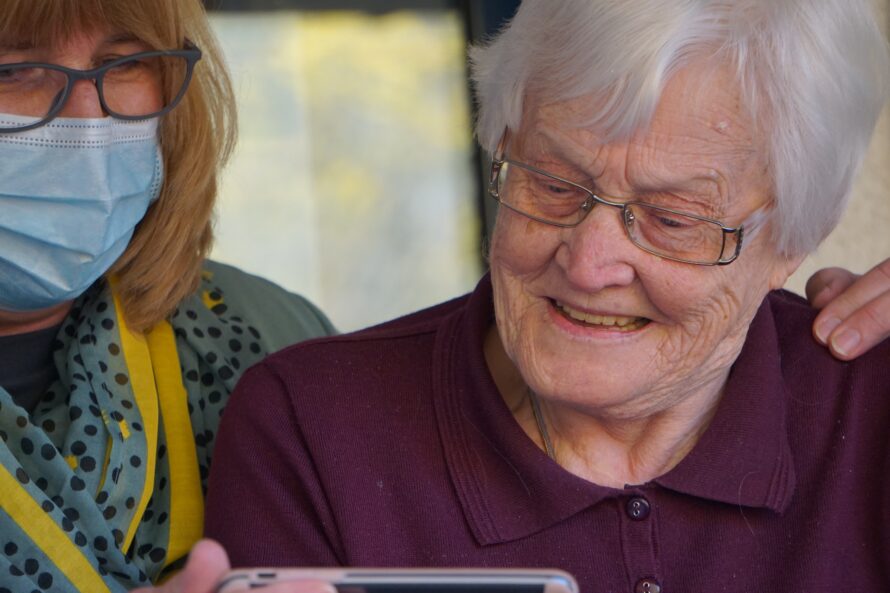 An elderly woman smiling and looking at a smartphone screen, accompanied by a person wearing a face mask and glasses, who is gently touching her shoulder.