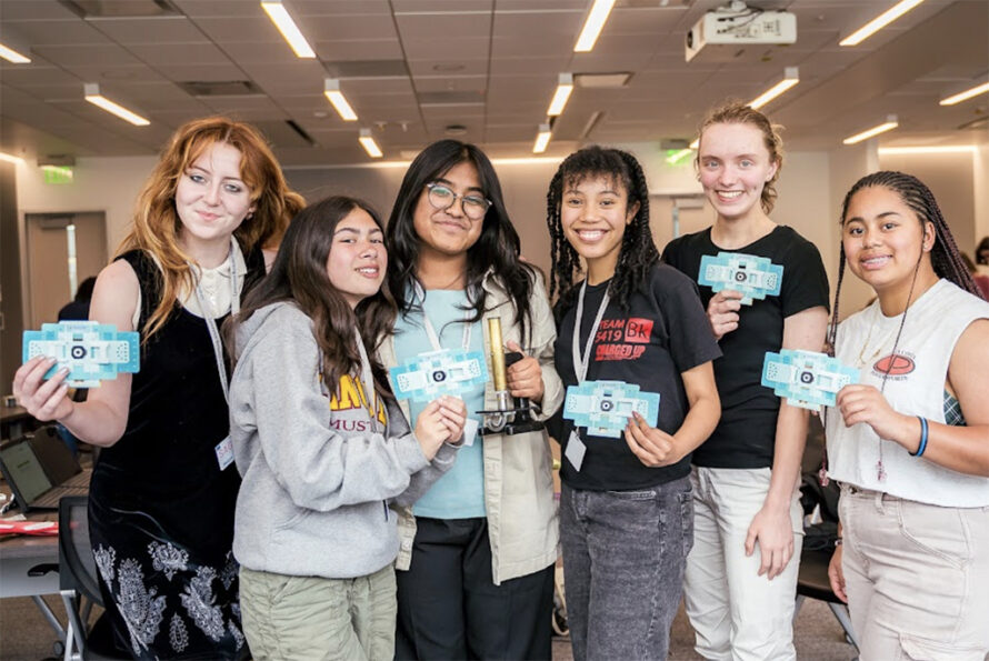 Group of diverse young women smiling and holding STEM-related cards, promoting the Girls Lead the Way STEM Summit happening on November 7