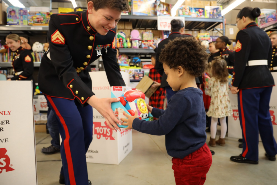 A US Marine, in uniform, hands a toy to a toddler in front of a wall of toys. Another Marine stands in the background.