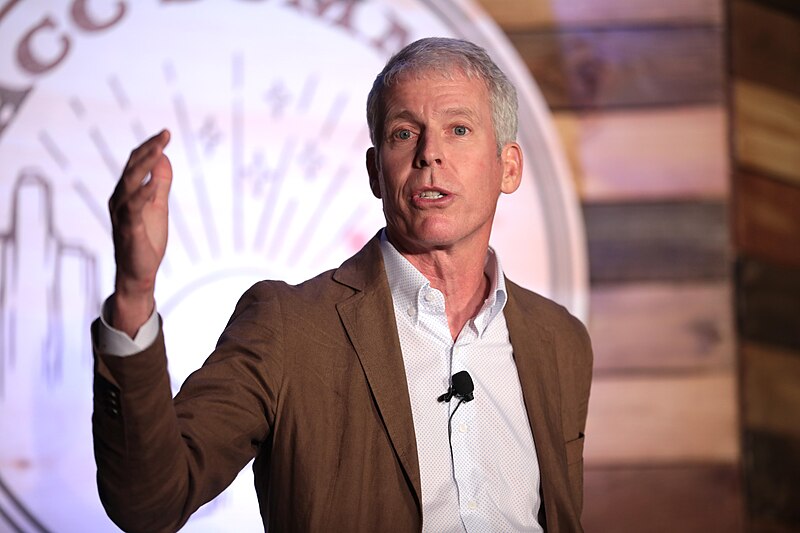 Person wearing a suit jacket with mic on lapel standing and gesturing with one hand.