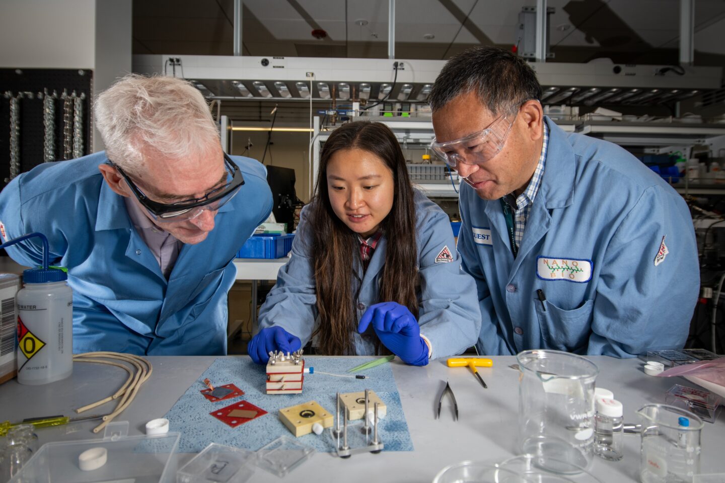 Photo of three persons in laboratory coats examining square device on top of table