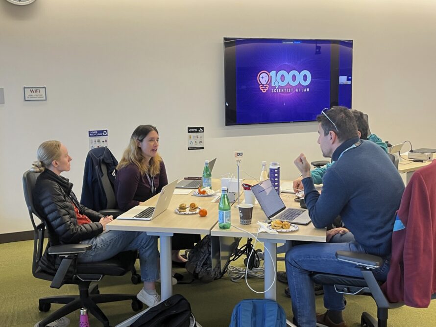 Four persons sitting at table with TV screen on the wall displaying the words "1000 Scientist AI Jam"