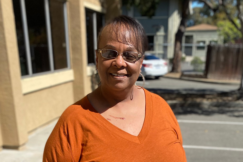 Daliscia Nunn, a person with dark brown hair tied back wearing sunglasses, hoop earrings, and an orange top, smiling photographed outdoors.