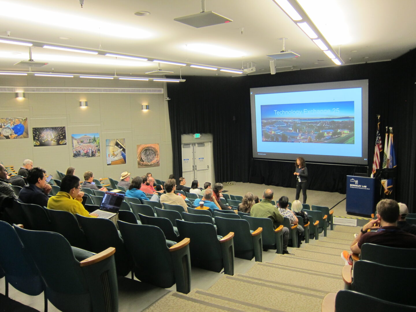 Auditorium with many people seated and listening to a speaker with large video screen in background.
