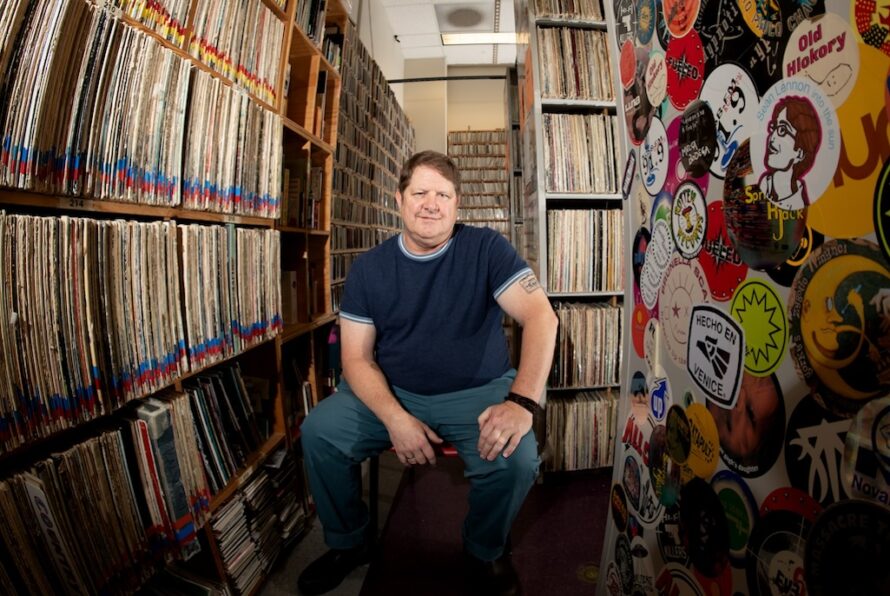 Person sitting on stool inside radio station with shelves of record albums on either side.