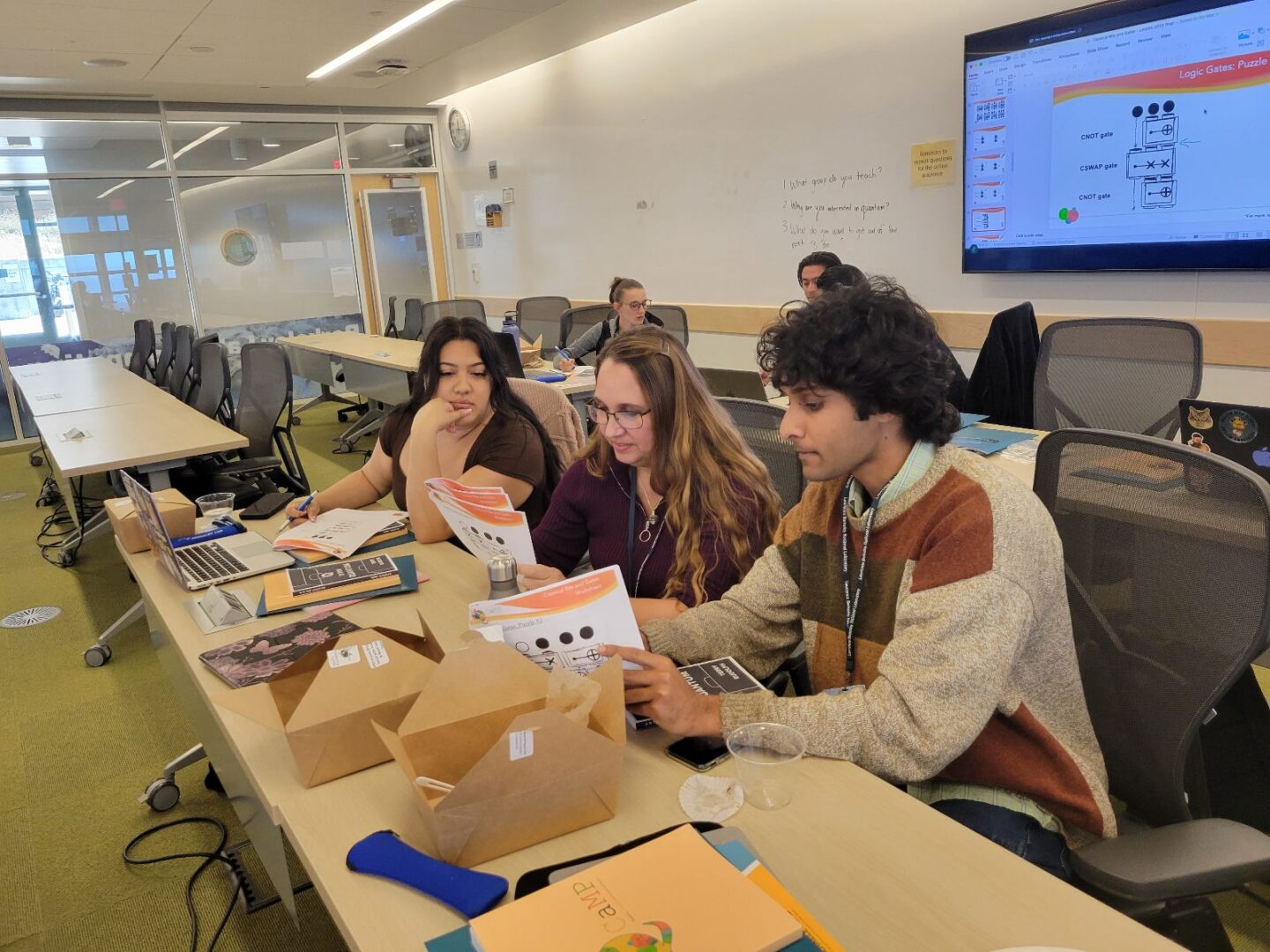 Three persons sitting at long table reading papers with wall-mounted monitor above them.