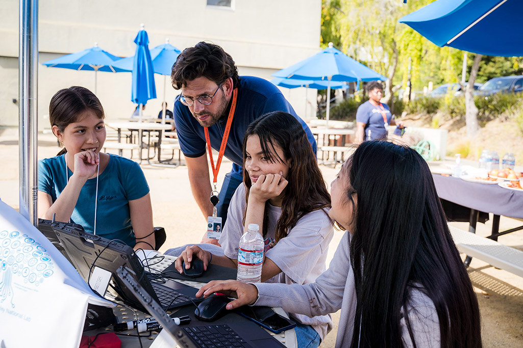 Adult reviewing information on a laptop with a group of students at an outdoor table.