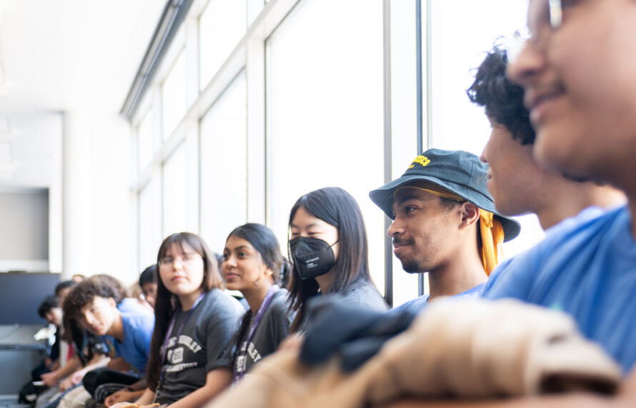 Group of persons sitting inside a room on a bench in front of large windows.