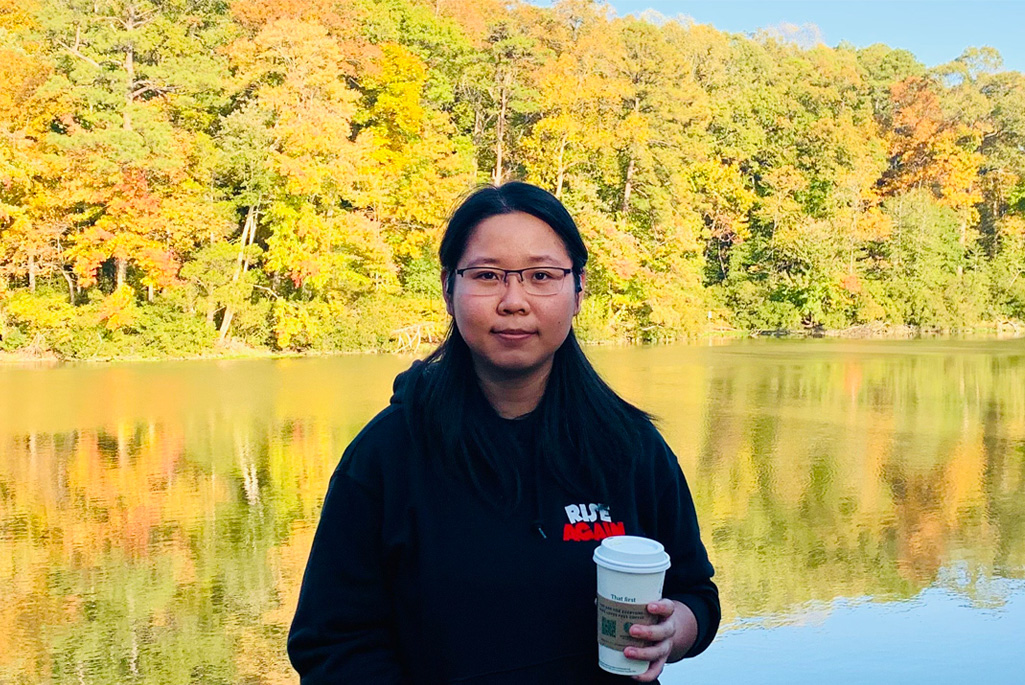 A person with medium-length dark hair wearing glasses and a black hoodie holding a coffee cup, standing by a lake with colorful autumn trees in the background.