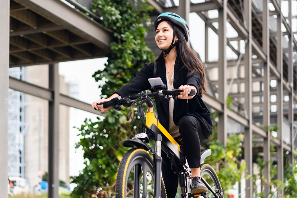 A person with long brown hair wearing a teal helmet, riding an electric bike to work in the city.