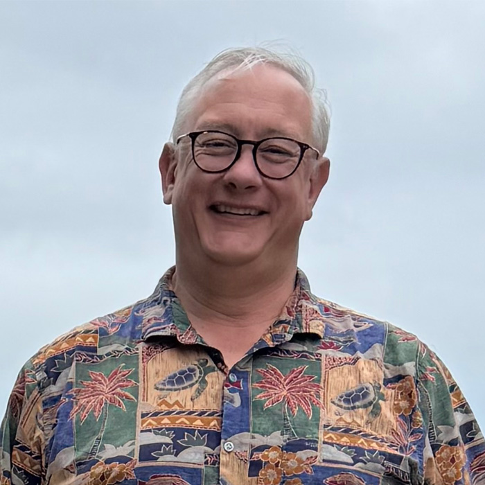 Bernard Hale, a person with short white hair wearing dark rimmed glasses and a colorful Hawaiian shirt, smiling outdoors.