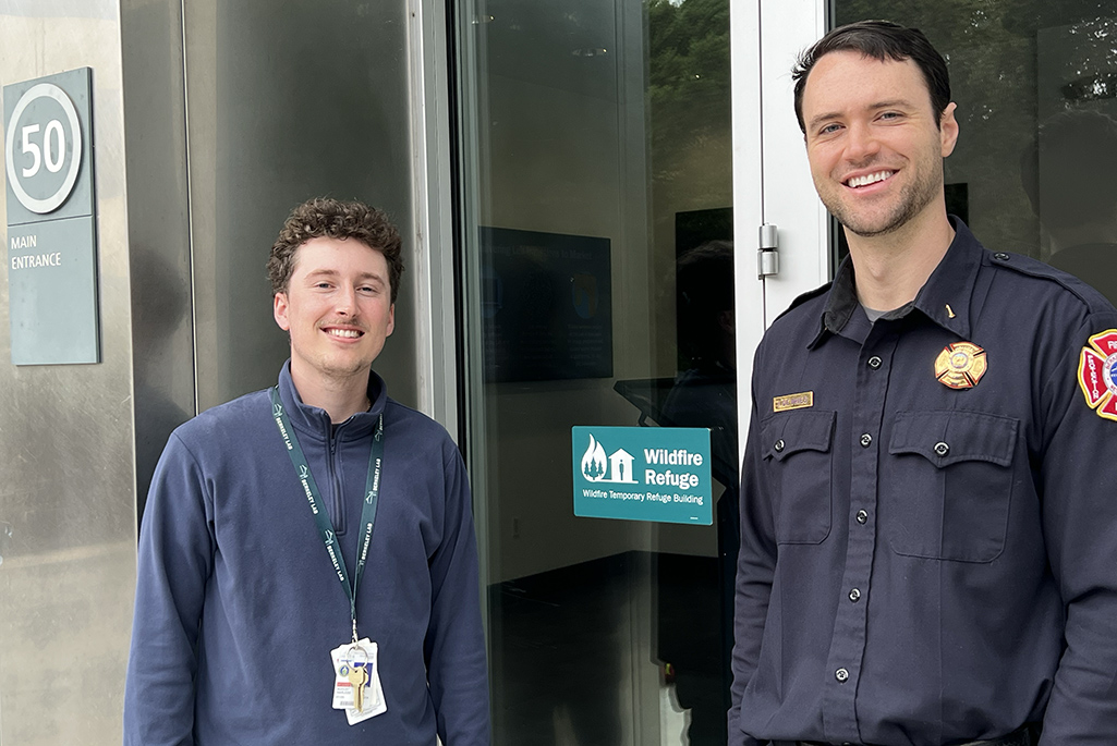 Two people, one in a fire uniform, outside a building standing next to sign on door.