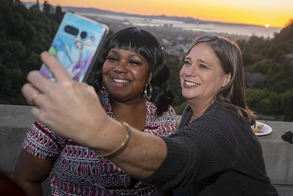 Two people standing outside with one holding a mobile phone to take photo with sunset in background