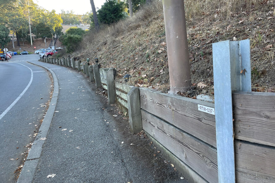 Roadside view with a sidewalk and wooden retaining wall on a sloped hillside. Metal beams and a utility pole line the side. Fallen leaves are scattered on the ground.