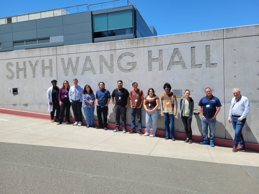 Persons standing in a line against a gray wall with the words Shyh Wang Hall.
