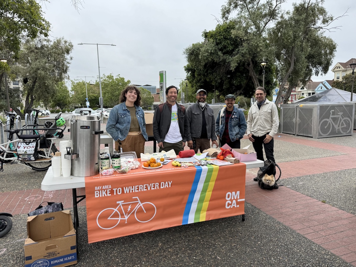 Five people stand outdoors behind a Bike to Wherever Day energizer booth from the OMCA. The booth has refreshments and snacks.