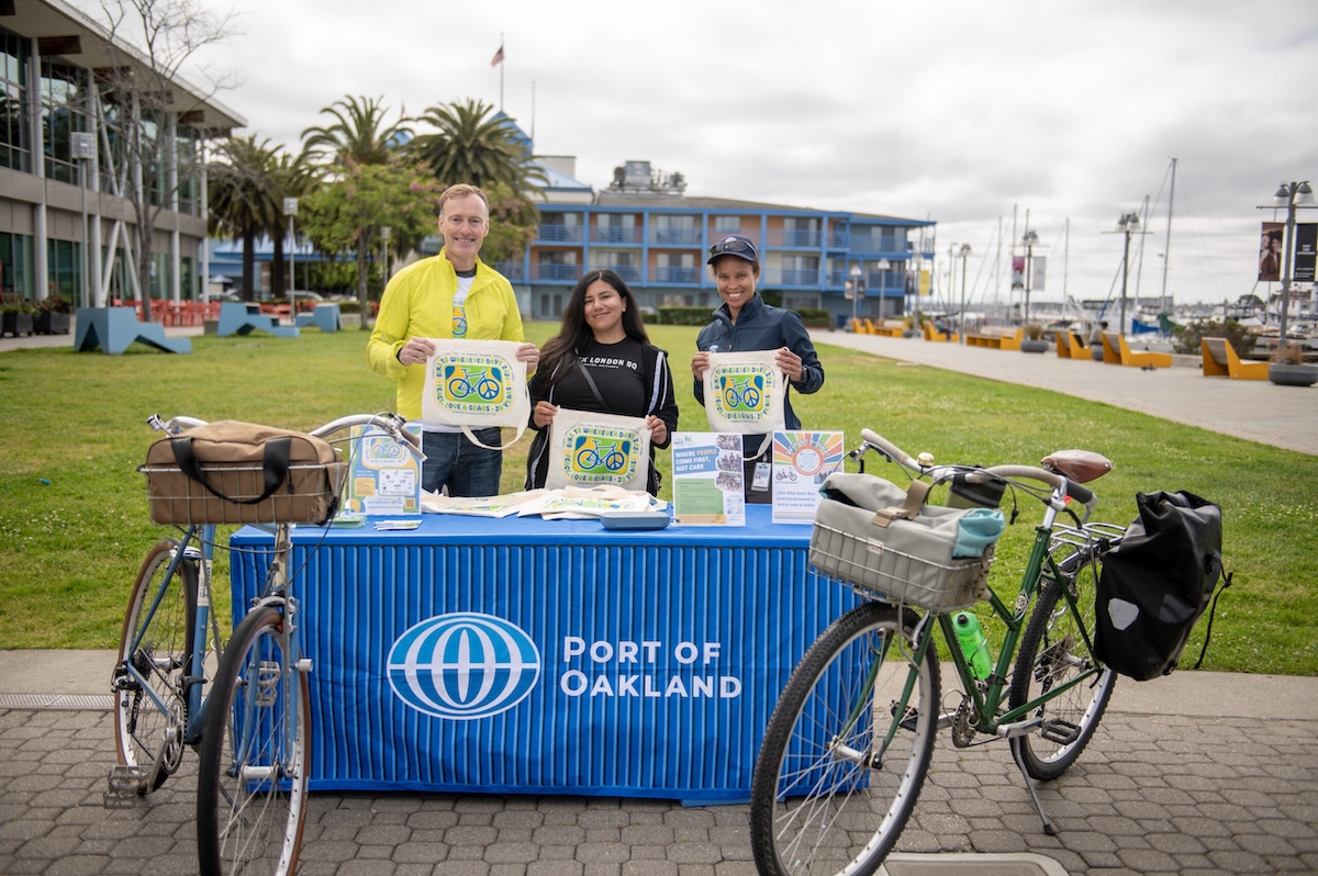Three people holding up Bike to Wherever Day tote bags at an energizer booth that has the Port of Oakland sign on ti. Two bikes flank the sides of their table.