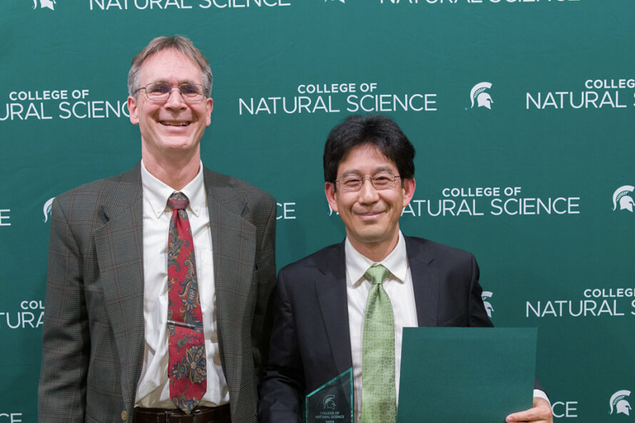 Two persons standing; one is holding an award and certificate of achievement.