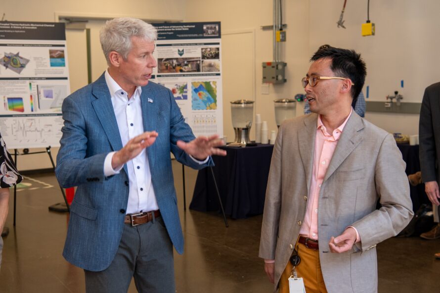Person in light blue jacket with hands raised to talk in discussion with other person with scientific equipment in background