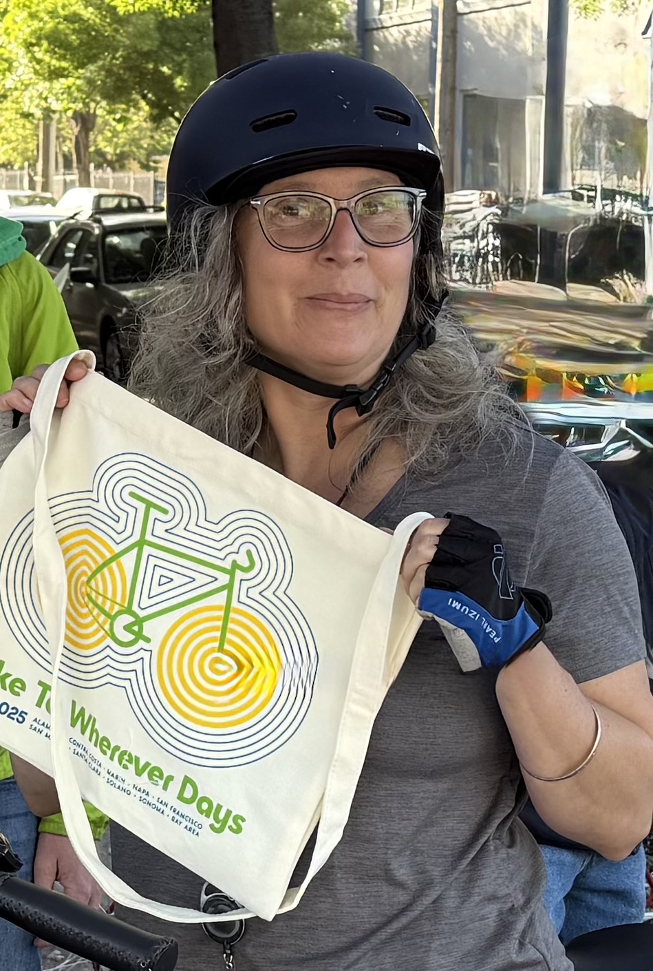 Person with long gray hair wearing a glasses and a bike helmet, holding up a tote bag featuring Bike to Wherever Days artwork.