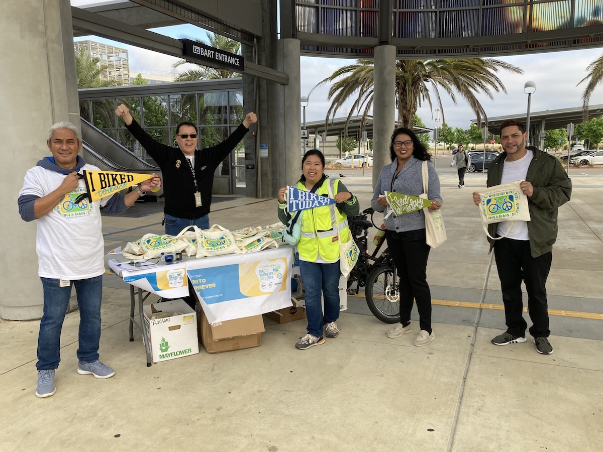 Five people smiling at a table set outdoors with Bike to Wherever Day tote bags. Three people are holding small flags that say, I biked today!, one person is holding the tote bag, and one person is raising both arms in a celebratory pose.