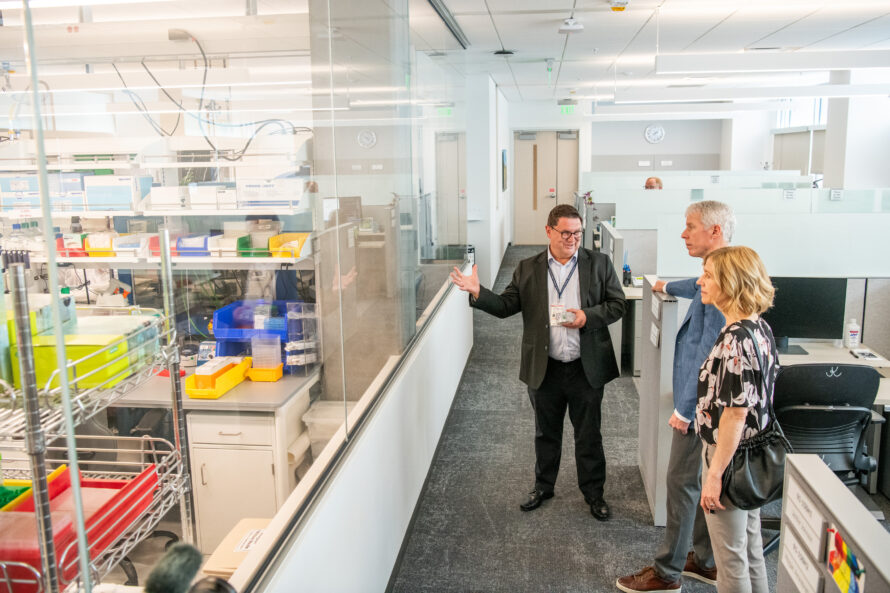 A person in dark suit jacket and pants gesturing toward scientific lab behind glass window as two people look on.