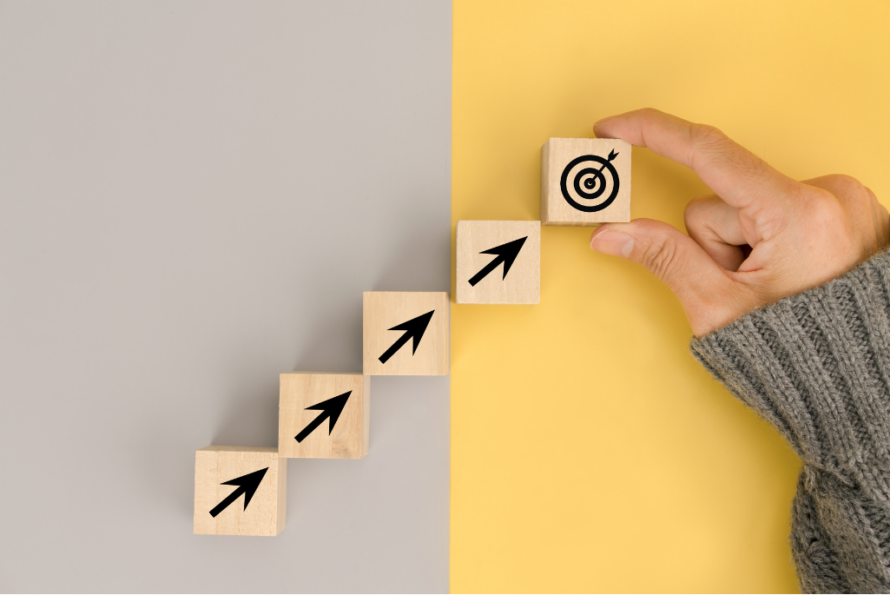 Image of four wooden blocks with up-arrows on each arranged in steps leading up to hand holding block with image of target.