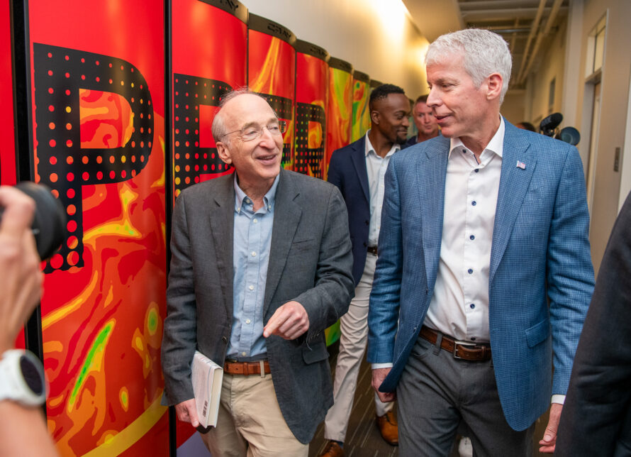 two person, both wearing suit jackets, talking as they walk past a supercomputer