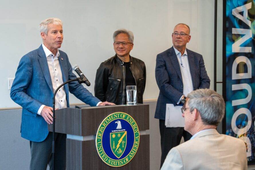 A person in a light blue jacket standing at a podium speaking into a microphone with two other people standing next to him.
