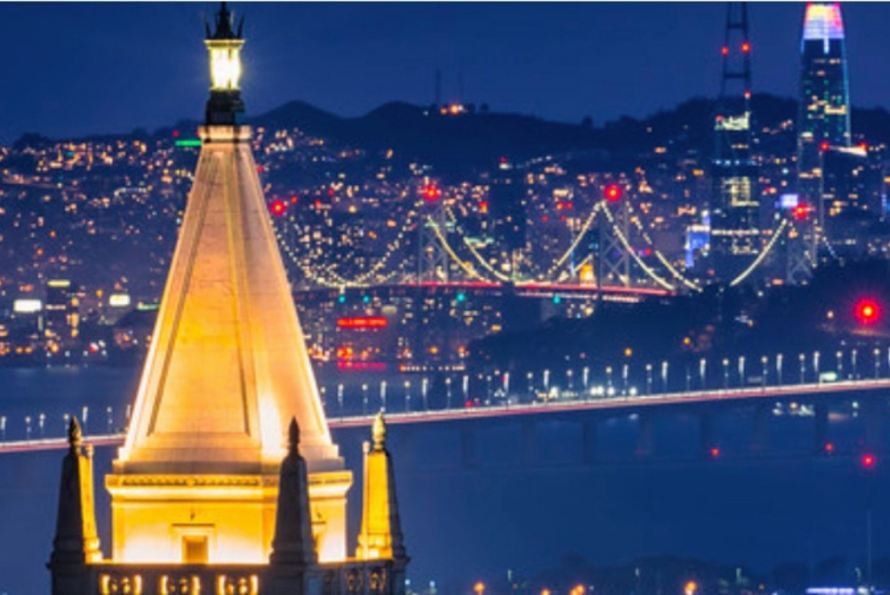 Night scene of UC Berkeley Carillon Tower with lit Bay Bridge in background.