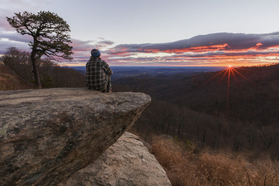 A person sits on a rocky ledge watching the sunset over rolling hills.