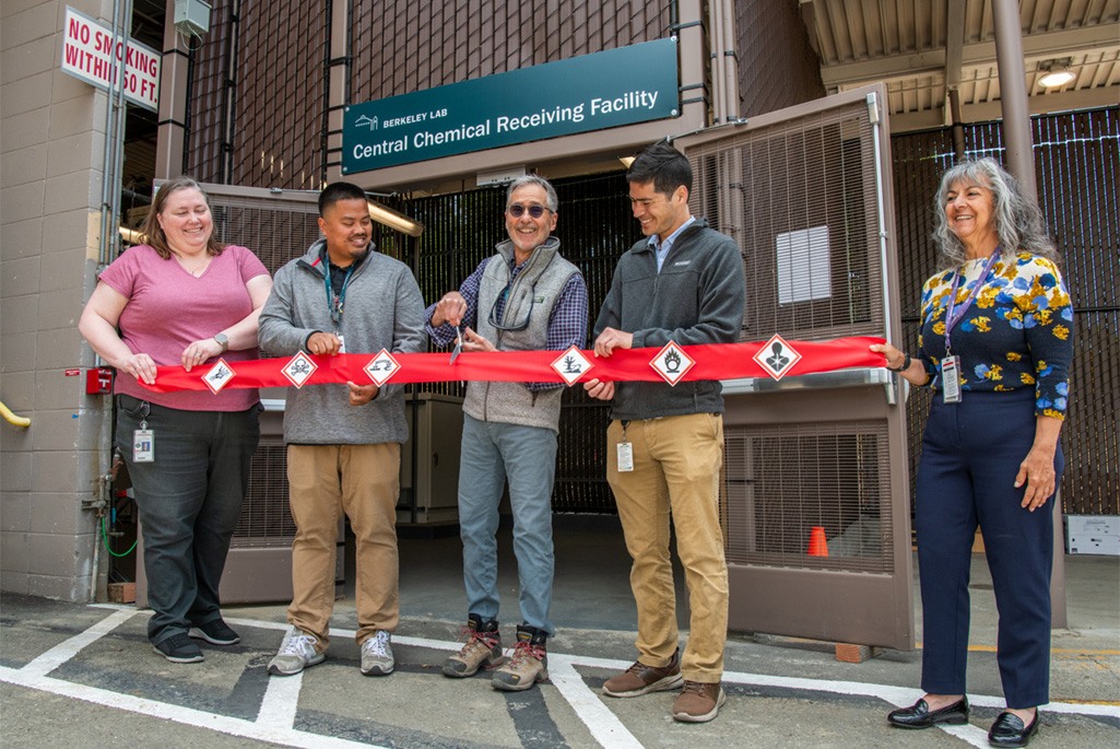Five individuals hold up a red ribbon with chemical safety pictograms attached for a ribbon-cutting ceremony.