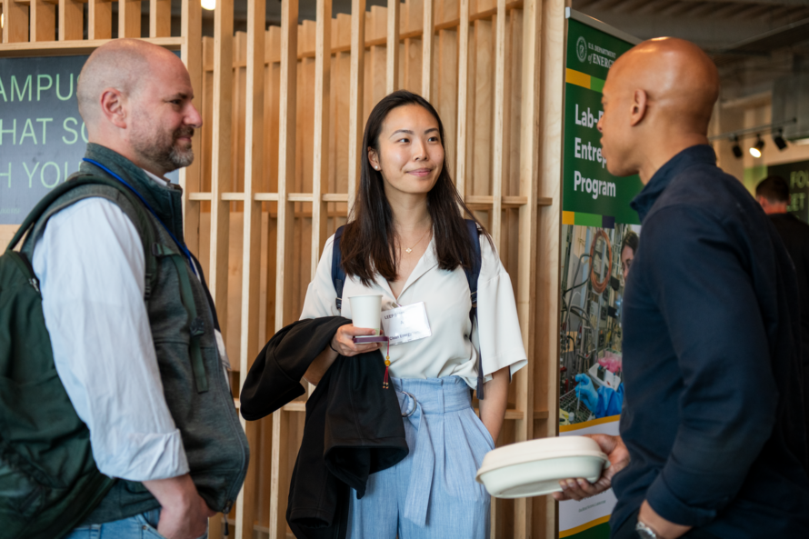 Three persons standing by booth inside hall engaged in discussion.