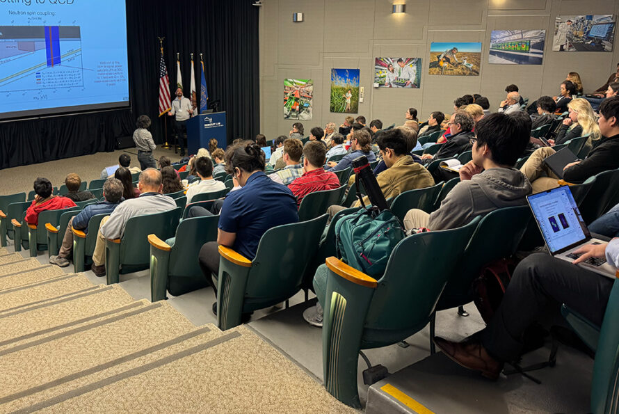 Large gathering of persons inside auditorium looking at wall-mounted monitor.