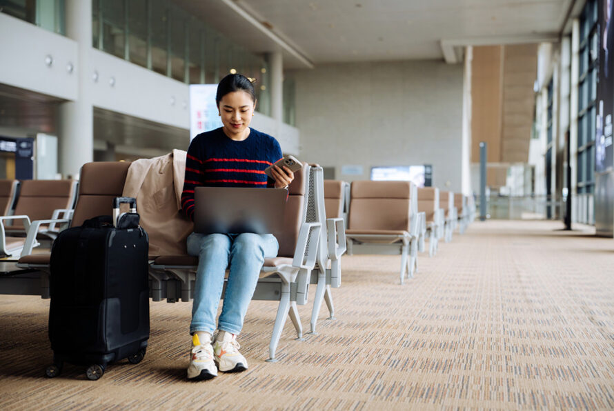 Person sitting at airport gate with suitcase next to them using a laptop and cell phone