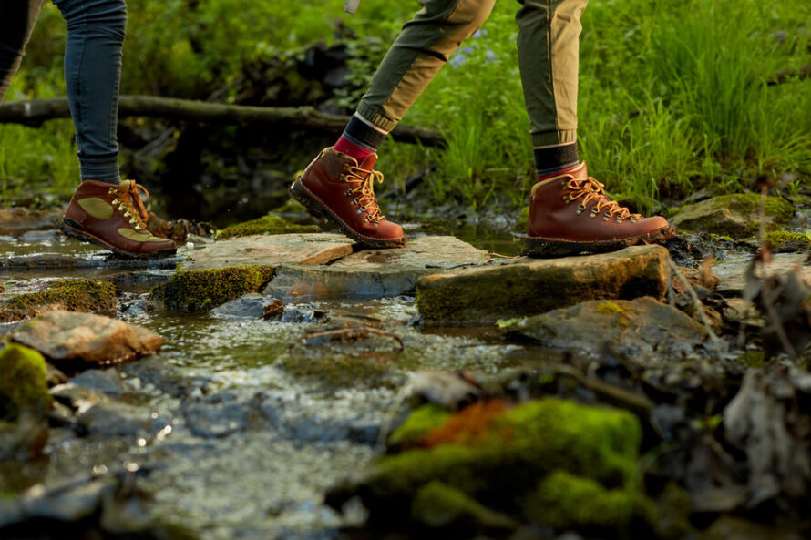 Closeup of two person's legs wearing boots walking over stones in a stream.