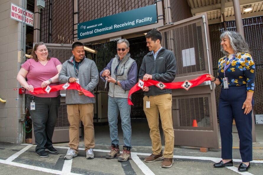 Five individuals hold up a red ribbon with chemical safety pictograms attached for a ribbon-cutting ceremony.