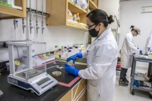 Person in chemical lab room wearing white lab coat and protective equipment.